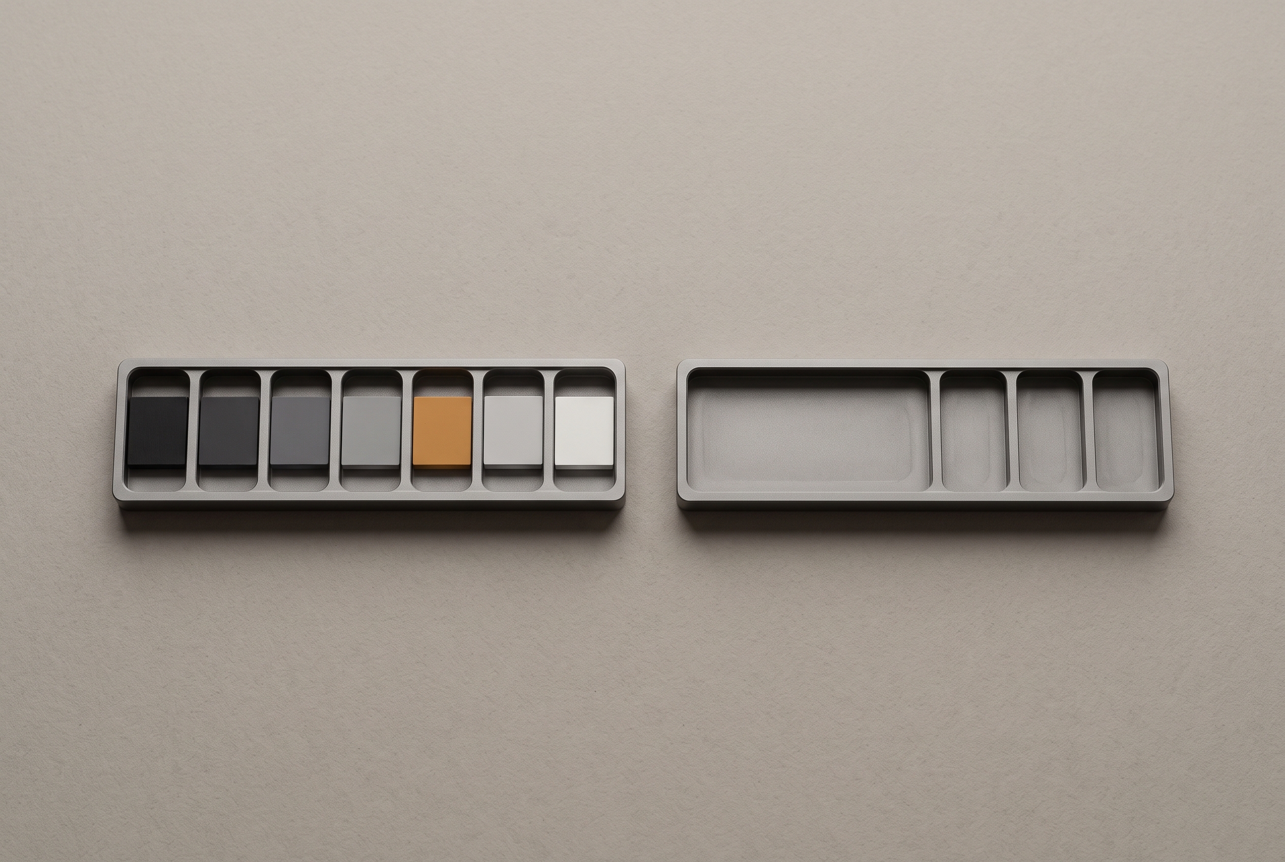 A row of small painted chips in a shallow tray, stepping from near-black to near-white, beside a second empty tray of the same shape.